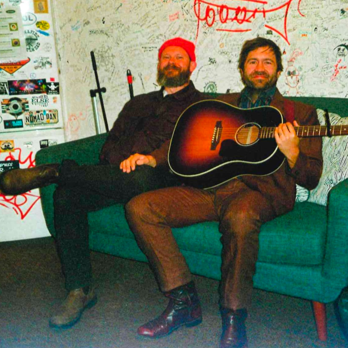 Bandmates sit on a green couch, holding guitars.