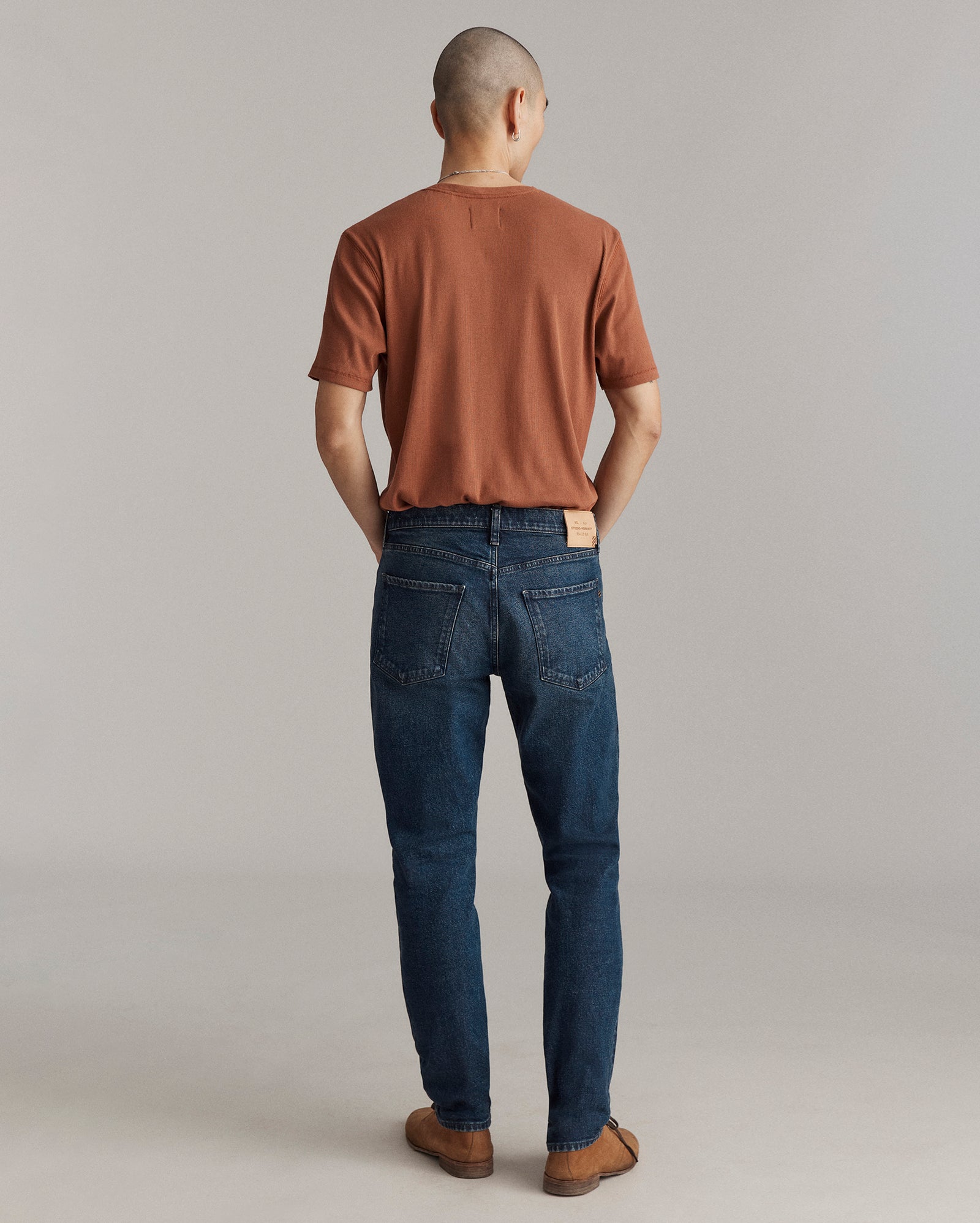A person stands facing away from the camera, wearing a brown short-sleeve shirt, blue jeans, and brown shoes, against a plain light gray background.