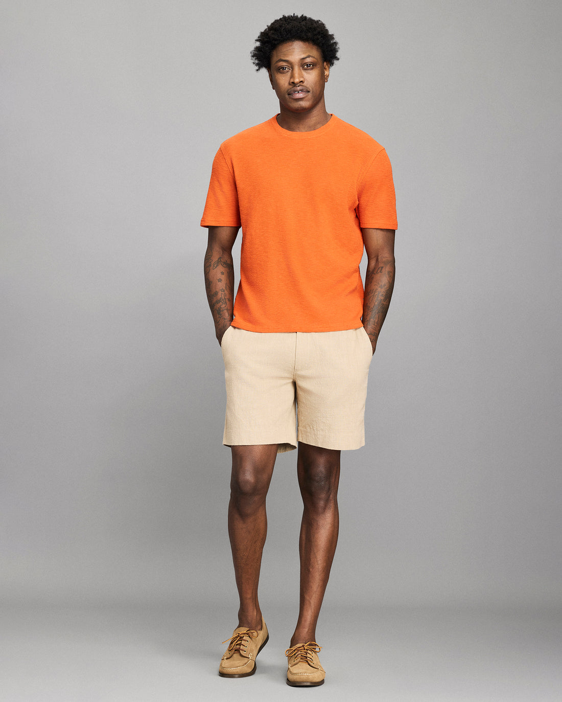 Man wearing an orange t-shirt and beige shorts against a gray background