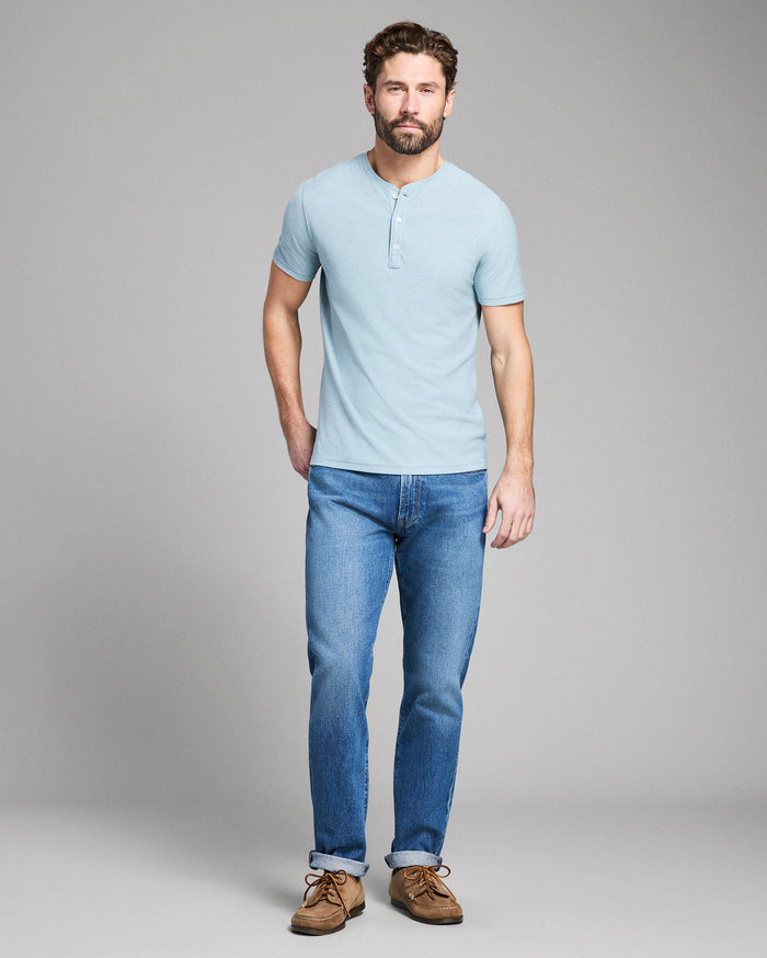 Man wearing a light blue henley shirt and jeans standing against a gray background