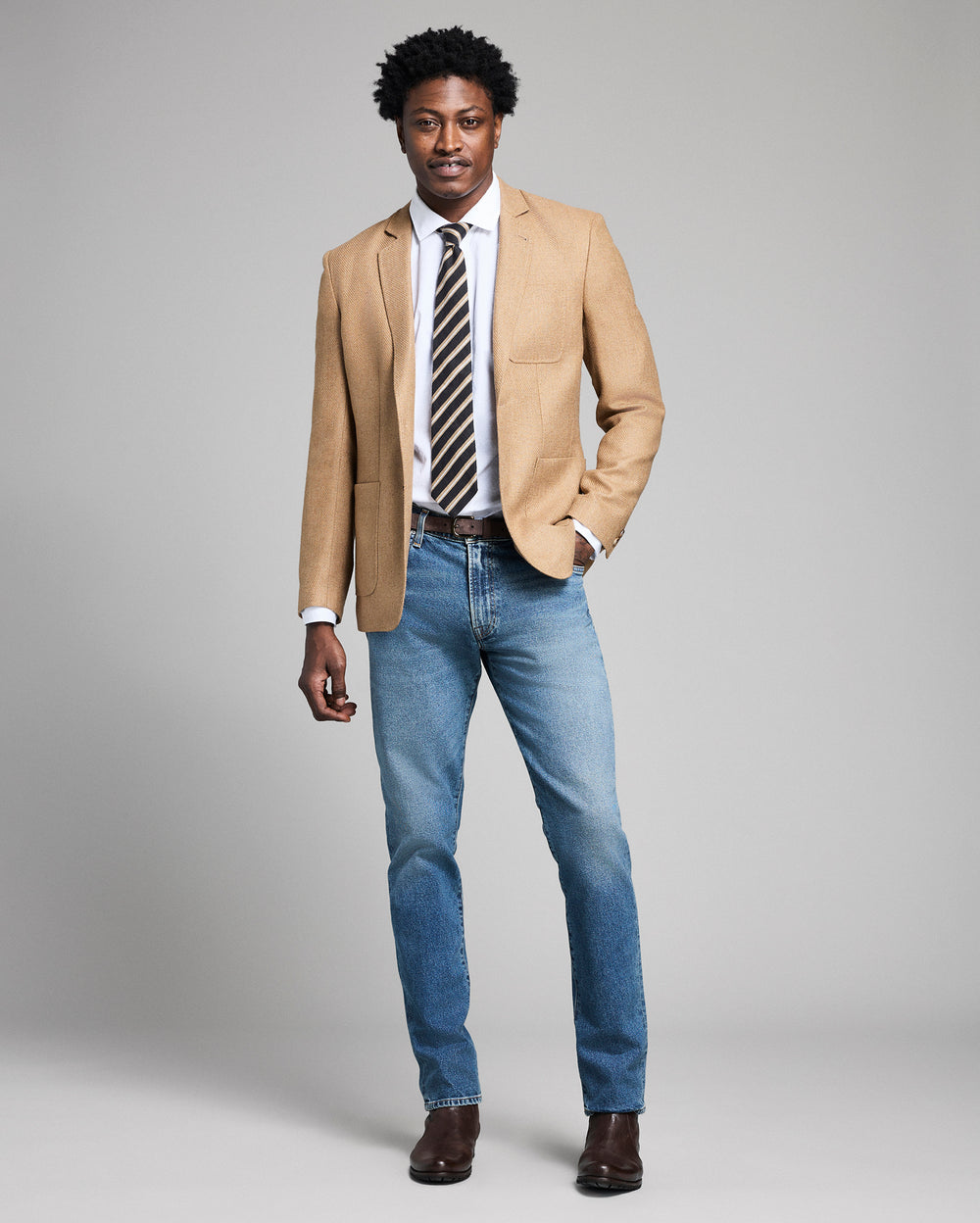 Man wearing a beige blazer, white shirt, striped tie, and blue jeans on a gray background