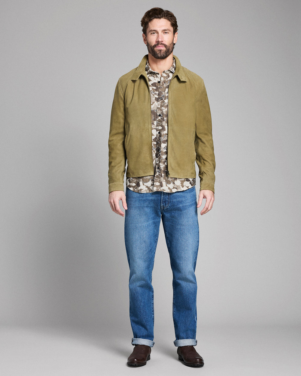 Man wearing a green jacket, camouflage shirt, and blue jeans on a gray background
