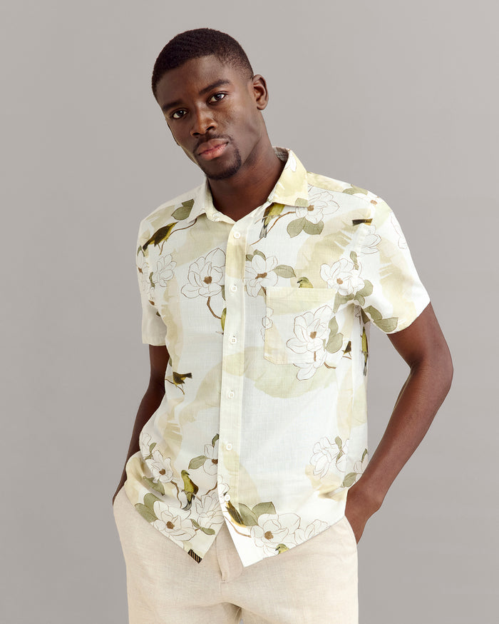 A man wearing a light-colored, short-sleeve button-up shirt with a floral pattern stands with his hands in his pockets against a plain gray background.