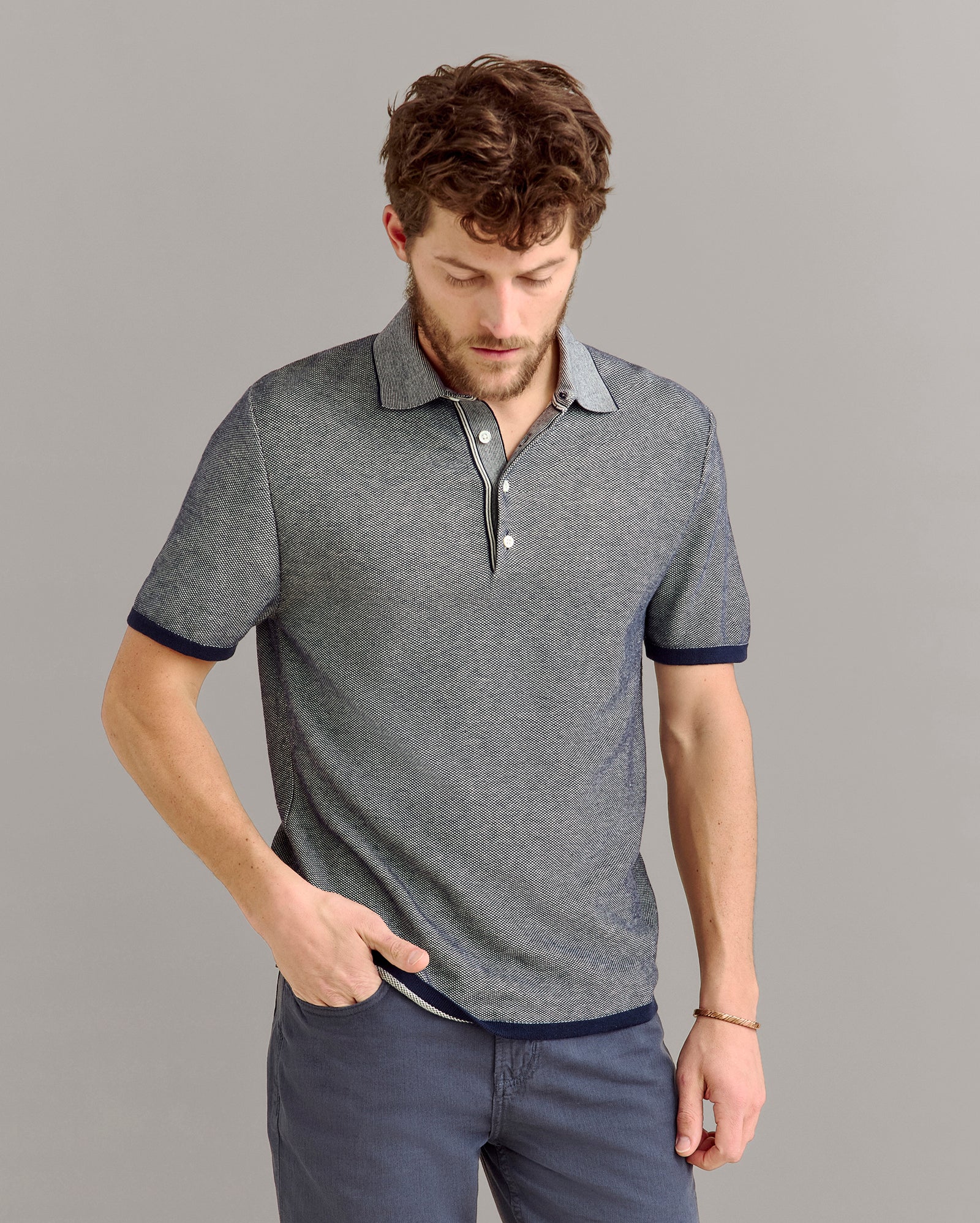 A man with curly brown hair and a beard is looking down while wearing a short-sleeved, gray collared polo shirt and dark gray pants. One hand is in his pocket against a plain light gray background.