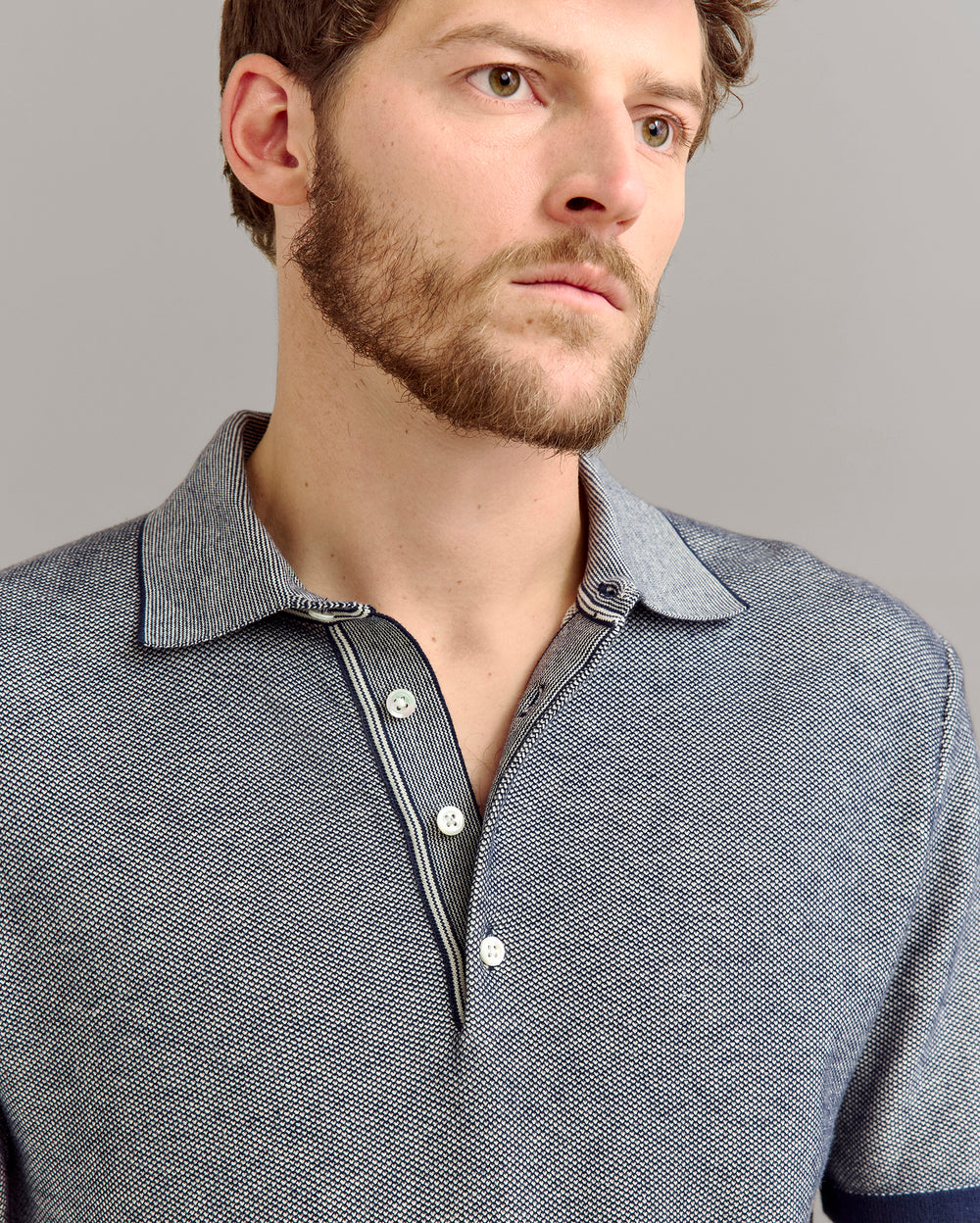 A man with a short beard and mustache wears a blue textured collared shirt partially unbuttoned, looking slightly to the side against a plain gray background.