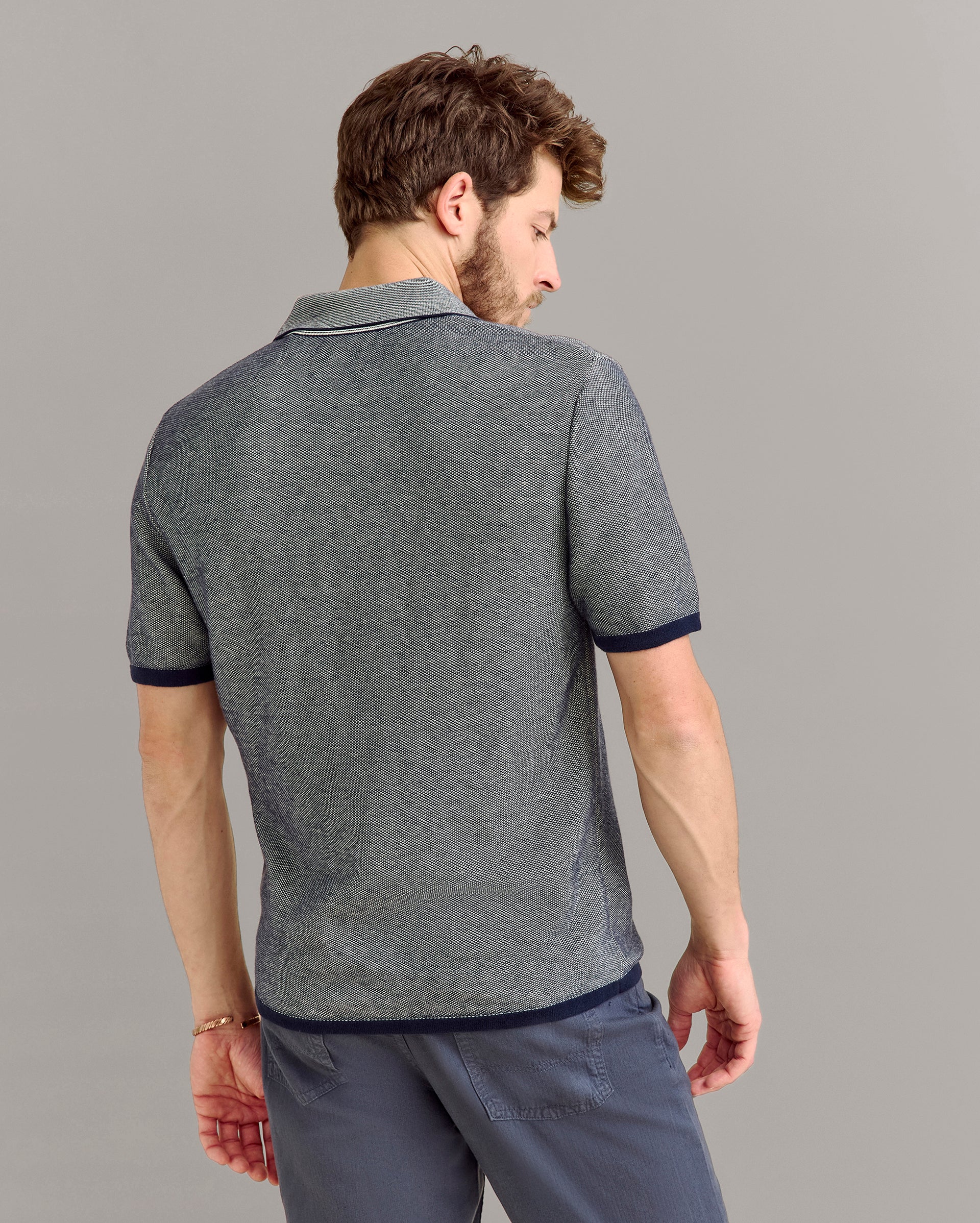 A man with short brown hair is standing and facing away, wearing a grey short-sleeve polo shirt with dark trim and blue pants, against a plain grey background.