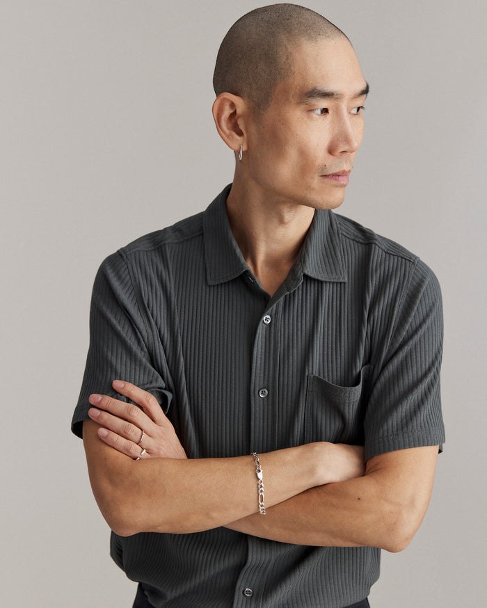 A person with a shaved head, wearing a short-sleeve, dark, pinstriped shirt and jewelry, stands with arms crossed and looks to the side against a plain background.
