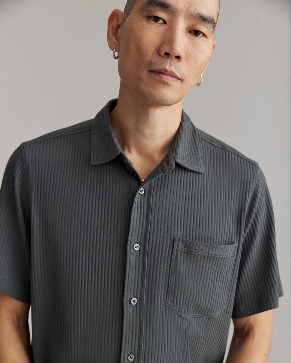 A person with short hair and hoop earrings wears a dark, short-sleeve, button-up shirt with vertical stripes and a chest pocket, standing against a neutral background.