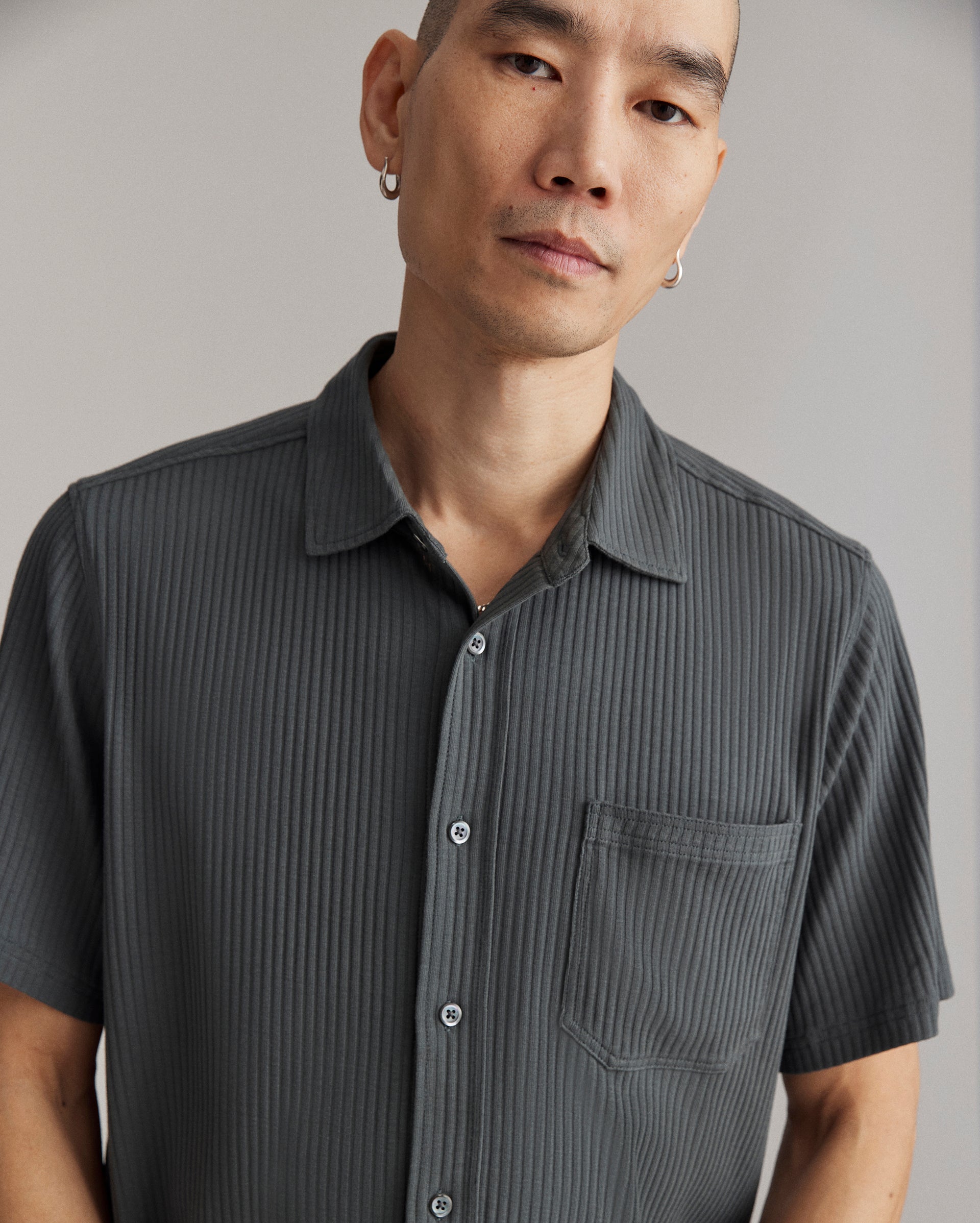 A person with short hair and hoop earrings wears a dark, short-sleeve, button-up shirt with vertical stripes and a chest pocket, standing against a neutral background.