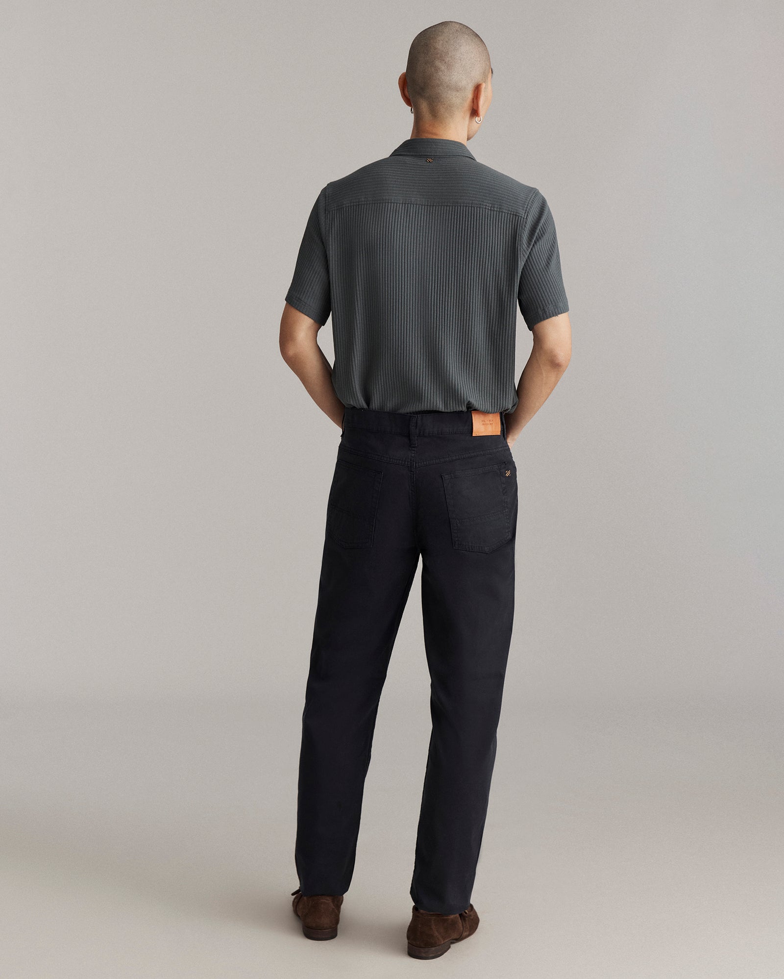 A person with short hair stands facing away, wearing a short-sleeve, dark gray shirt and black pants with a tan patch on the waistband, and brown shoes, against a plain light background.