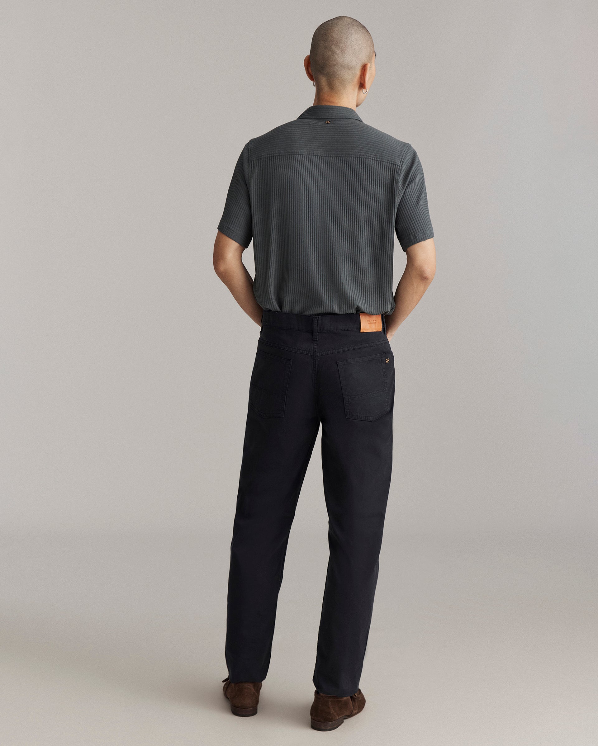 A person with short hair stands facing away, wearing a short-sleeve, dark gray shirt and black pants with a tan patch on the waistband, and brown shoes, against a plain light background.