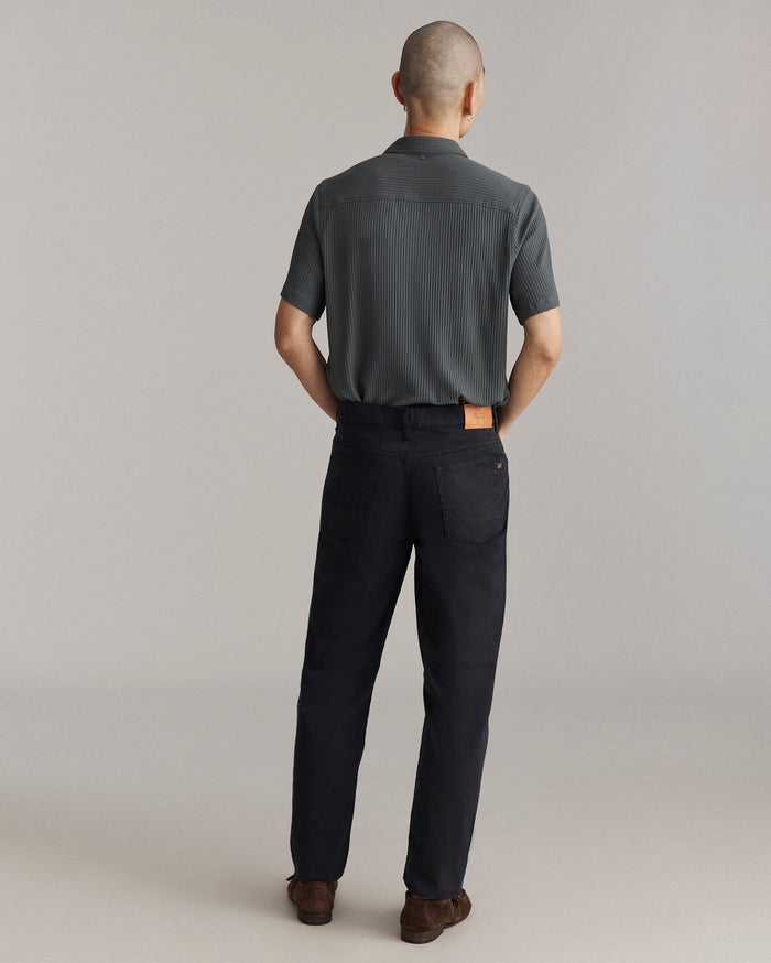 A person with short hair stands facing away, wearing a short-sleeve, dark gray shirt and black pants with a tan patch on the waistband, and brown shoes, against a plain light background.