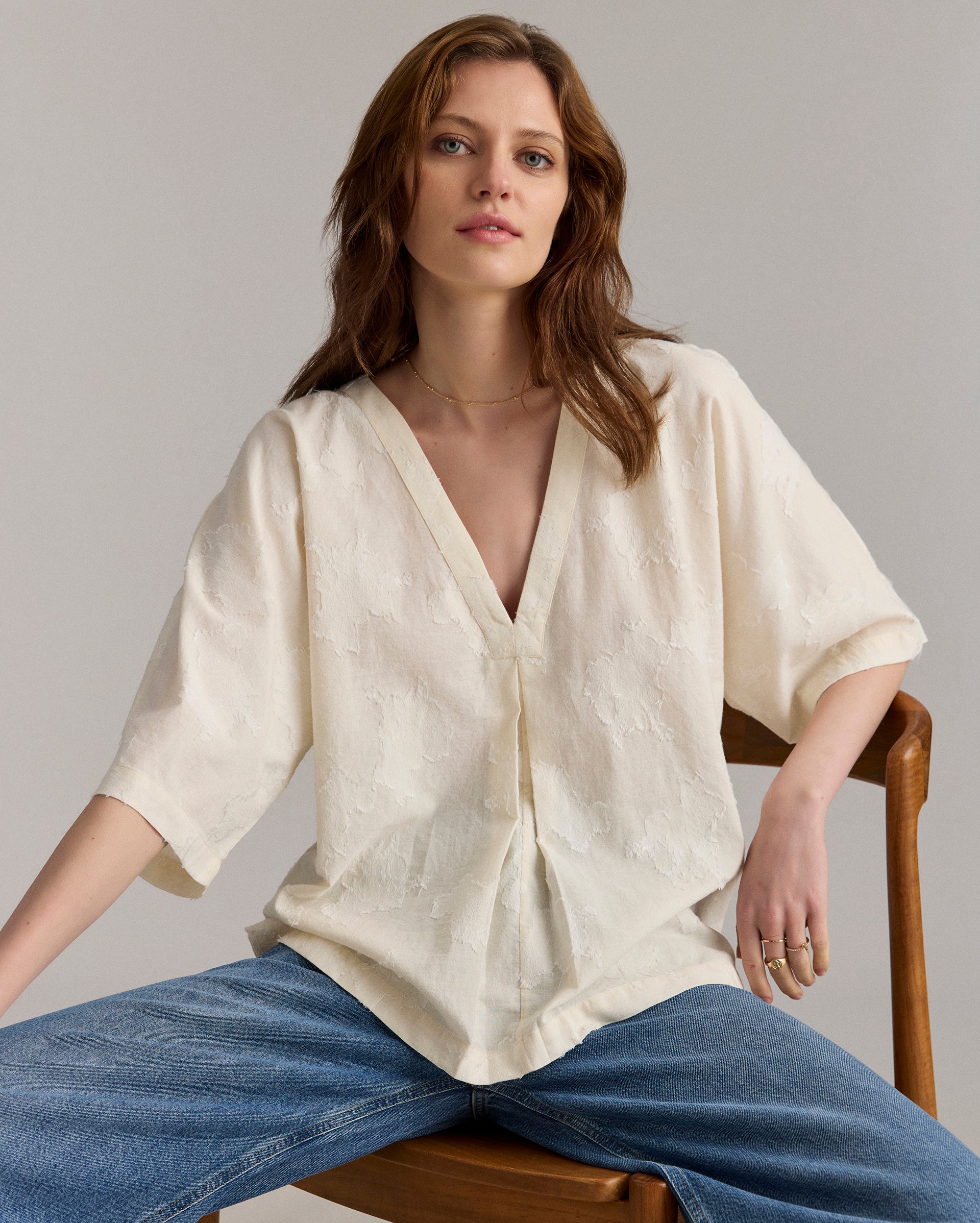 A woman with wavy brown hair sits on a wooden chair, wearing a loose, cream-colored V-neck blouse with subtle patterns and blue jeans. She looks relaxed, with one hand on her knee and the other resting on the chair.