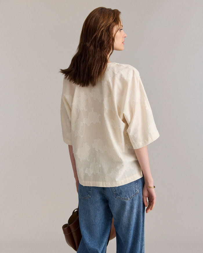 A woman with brown hair wearing a loose, cream-colored blouse with a floral pattern and blue jeans stands facing away, holding a brown handbag in her left hand against a neutral background.