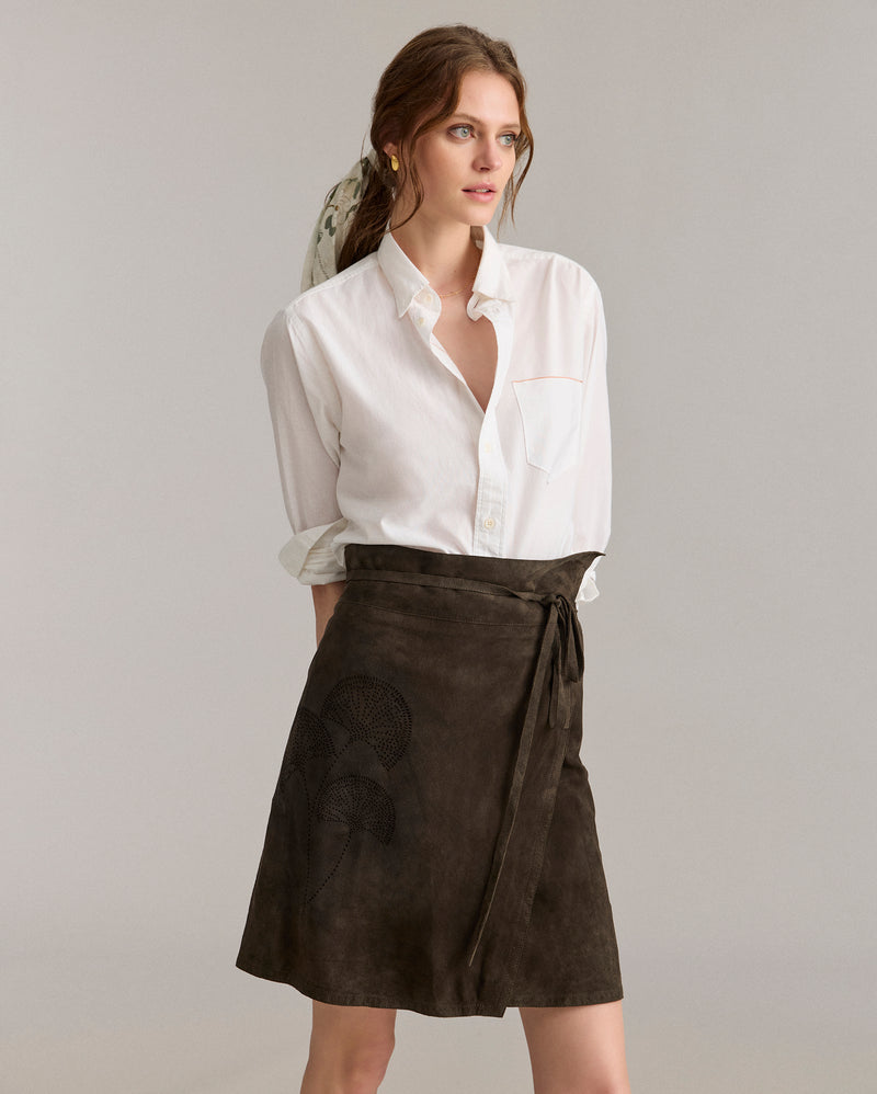 A woman wearing a white button-up shirt with rolled sleeves and a dark brown wrap skirt stands against a plain, light background, looking to the side with a neutral expression. Alternate