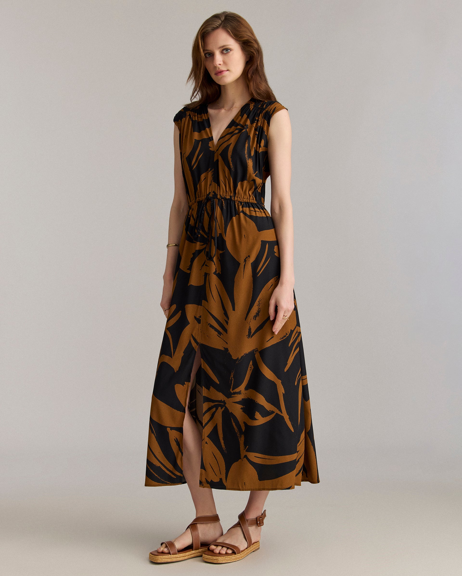 Woman wearing a sleeveless, black and brown floral maxi dress with a v-neck and front slit, paired with tan wedge sandals, standing against a plain light background.