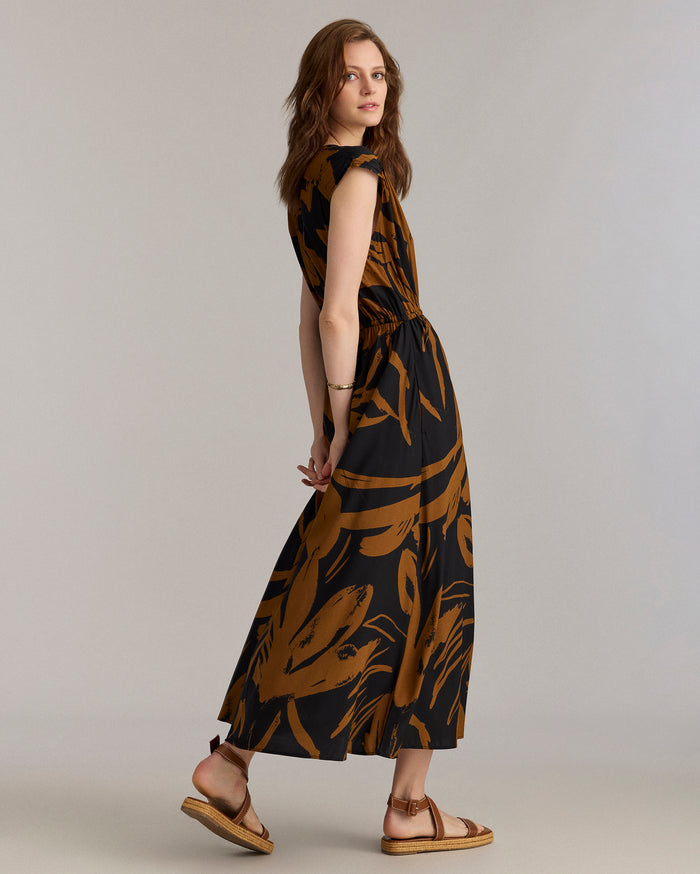 A woman with light brown hair wears a black and brown patterned maxi dress and tan sandals, standing sideways and looking over her shoulder against a plain light background.