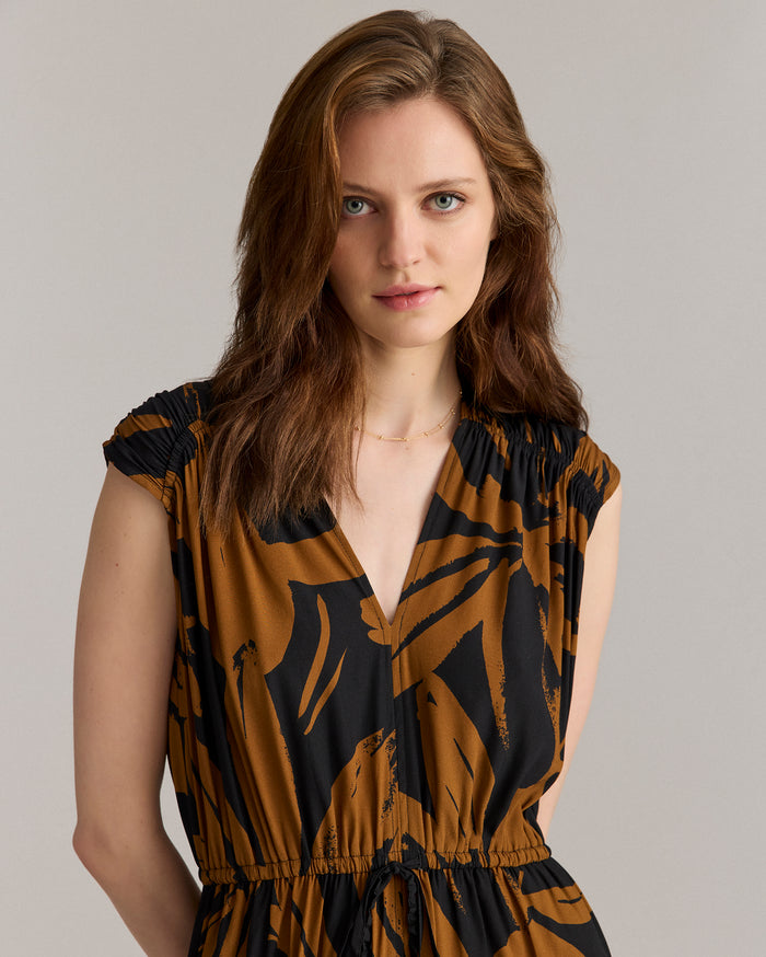 A woman with light skin and wavy brown hair stands against a plain background, wearing a short-sleeved brown and black patterned dress with a V-neckline and a subtle gold necklace. She is looking at the camera.