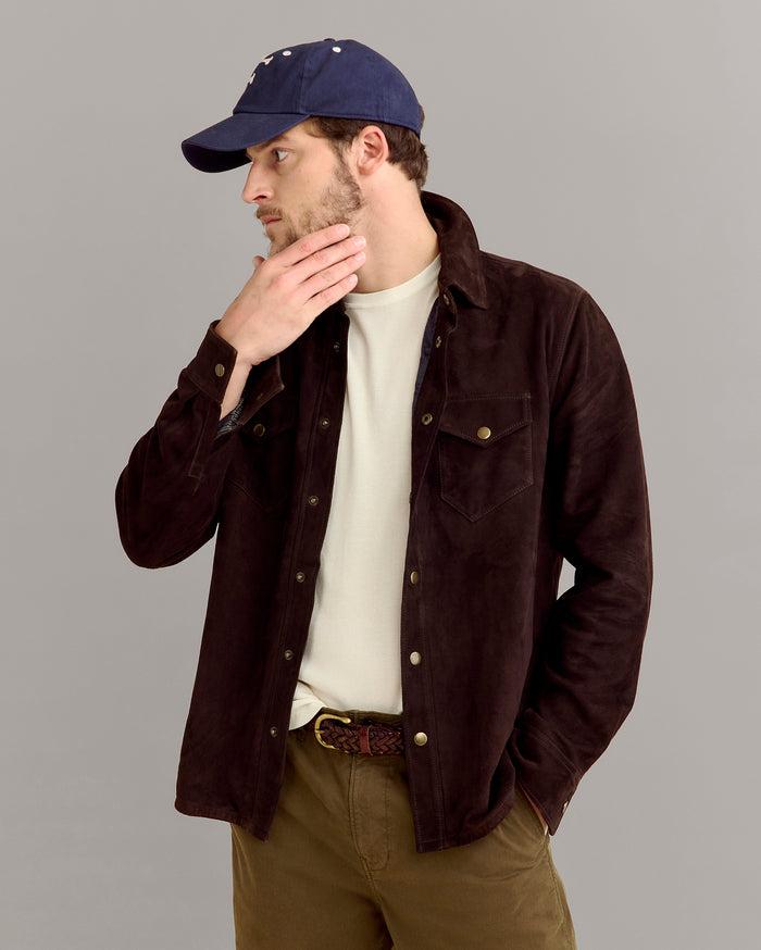 A man wearing a blue baseball cap, off-white shirt, dark brown jacket, and olive green pants stands against a plain gray background, looking to the side with one hand touching his chin.