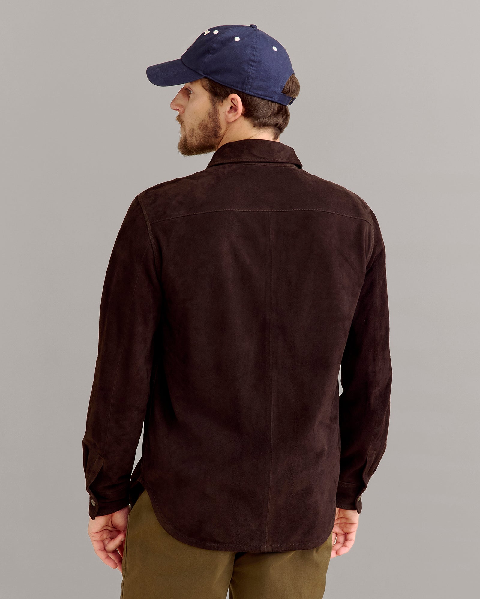 A man wearing a dark brown button-up shirt, olive green pants, and a navy blue baseball cap is standing and facing away from the camera against a plain light gray background.