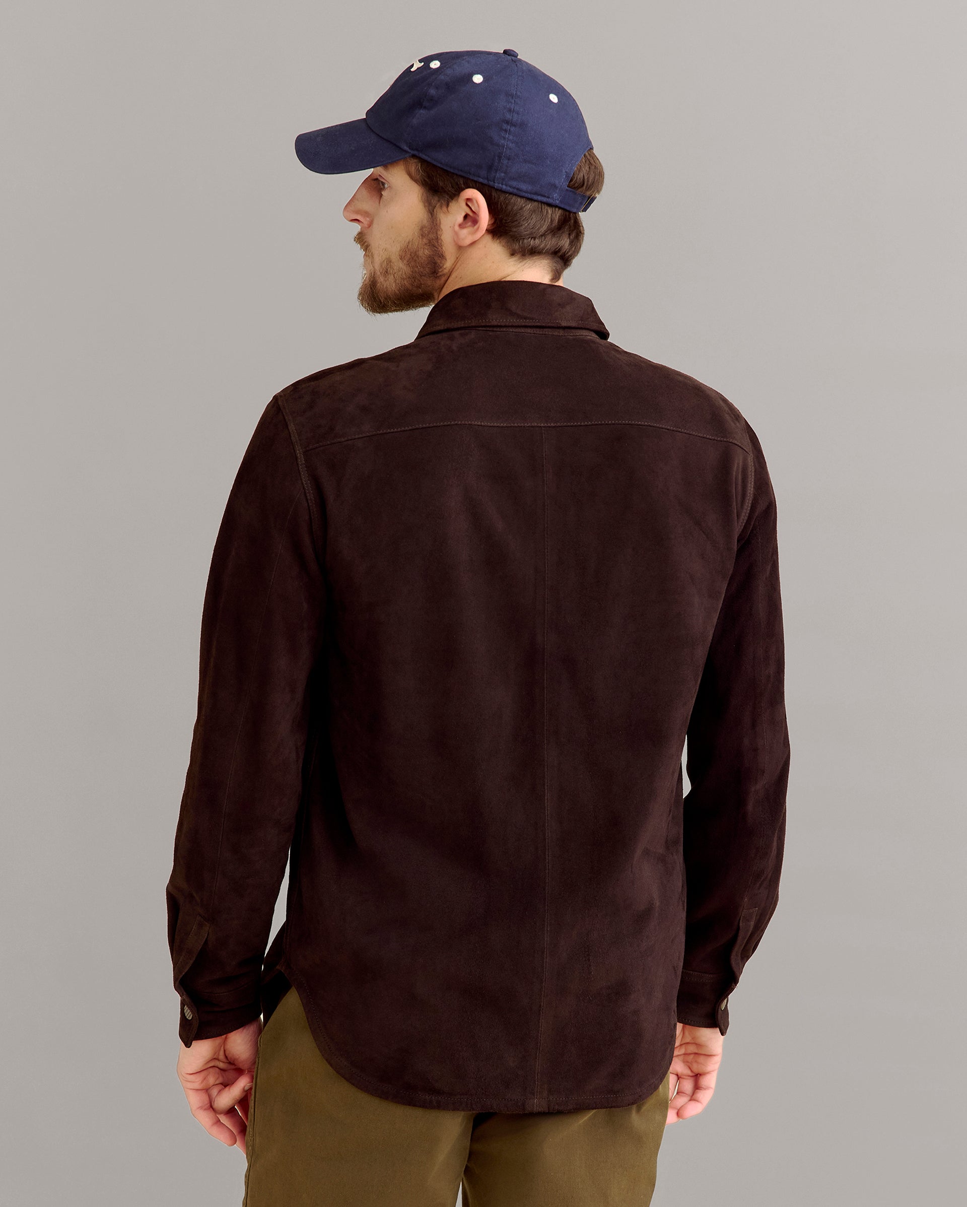 A man wearing a dark brown button-up shirt, olive green pants, and a navy blue baseball cap is standing and facing away from the camera against a plain light gray background.
