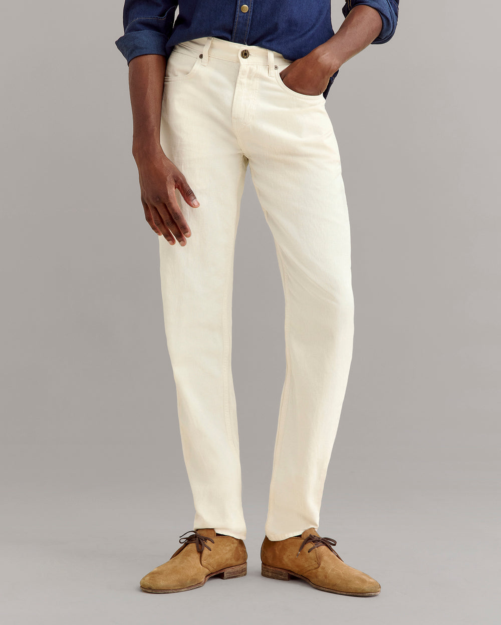 A person wearing cream-colored jeans, a dark blue button-up shirt, and tan suede shoes stands against a plain gray background, with one hand in their pocket.
