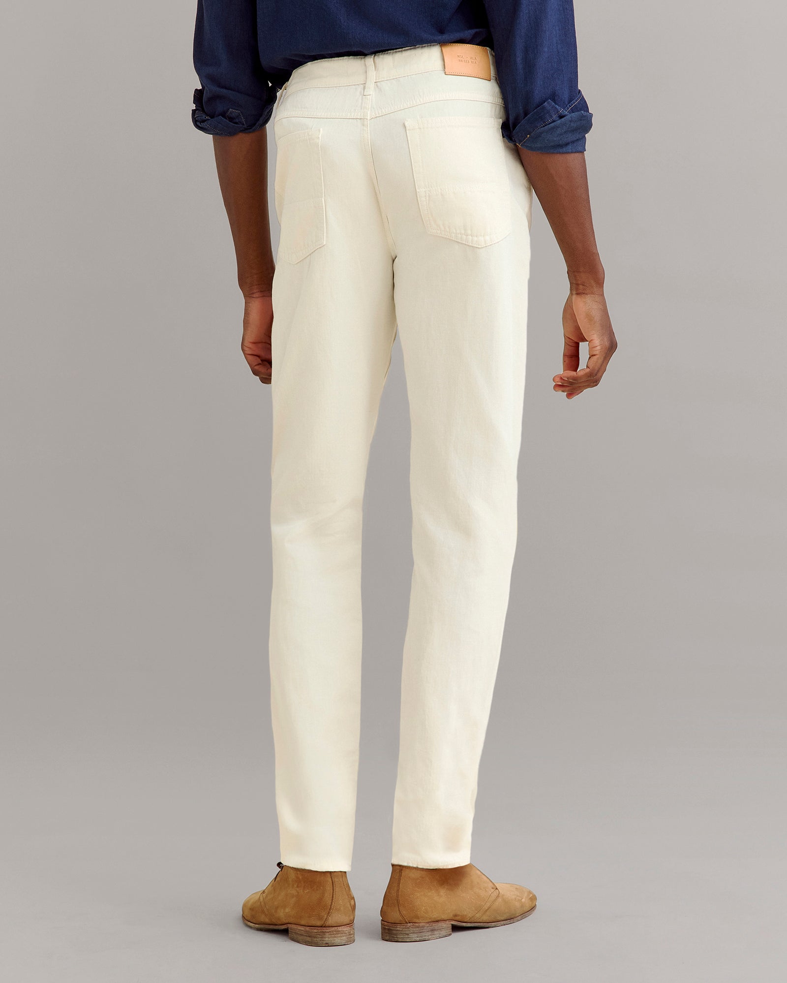 A person stands facing away, wearing a dark blue shirt, light cream jeans, and tan suede shoes, against a plain gray background.