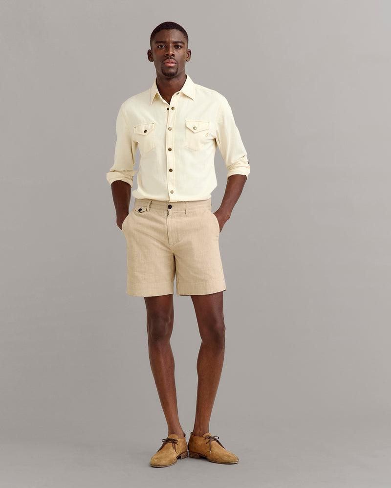 A man stands against a plain gray background wearing a light yellow button-up shirt with rolled-up sleeves, beige shorts, and tan shoes. His hands are in his pockets and he looks forward. Alternate