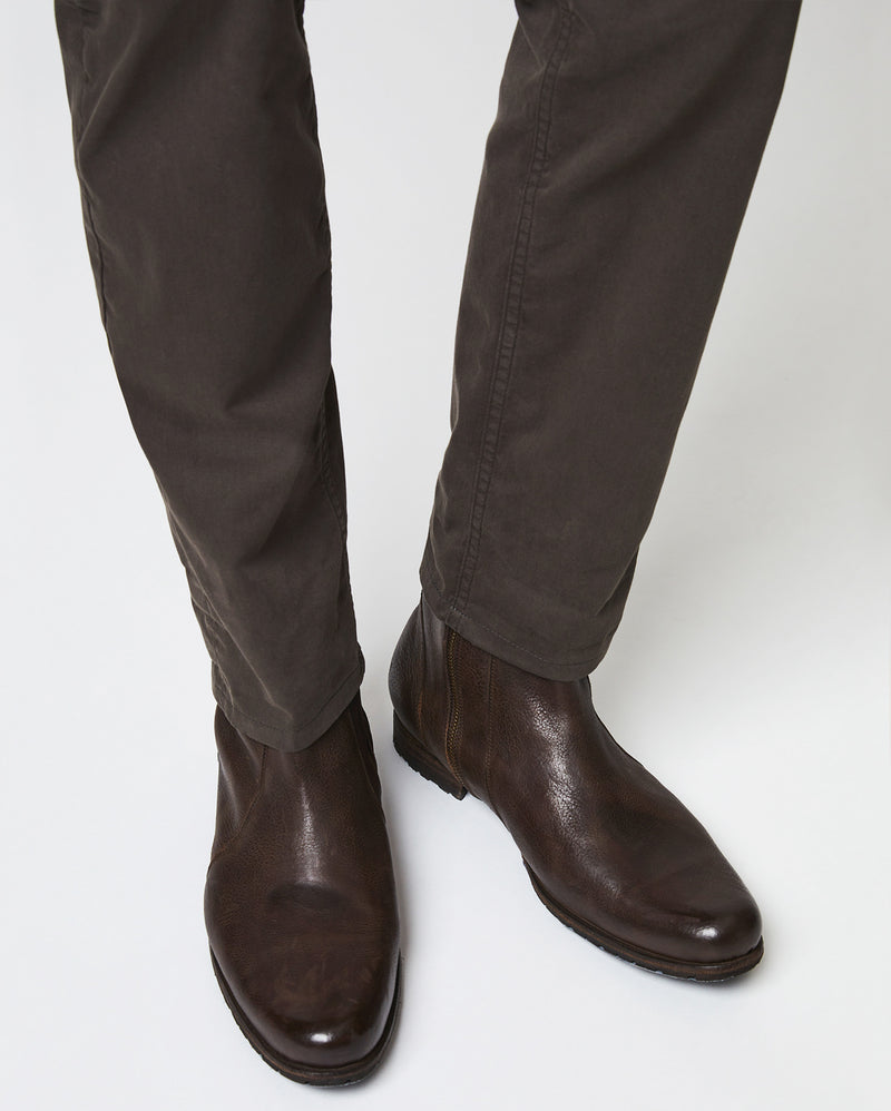 A person wearing dark brown leather Chelsea boots and dark brown pants standing on a light-colored surface. Alternate