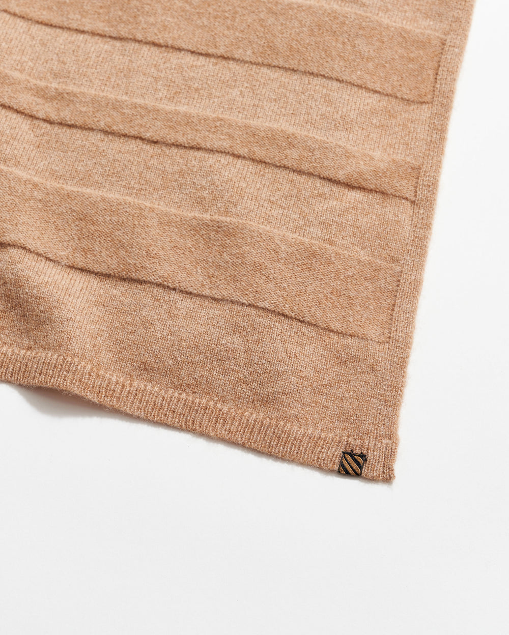 A close-up of a light brown knitted fabric with horizontal textured stripes, finished with a ribbed edge and a small rectangular tag in the corner on a white background.