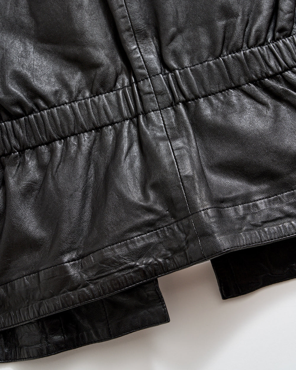 Close-up of a black leather garment, showing gathered and stitched detailing along the waist and hem areas, highlighting the texture and sheen of the material.