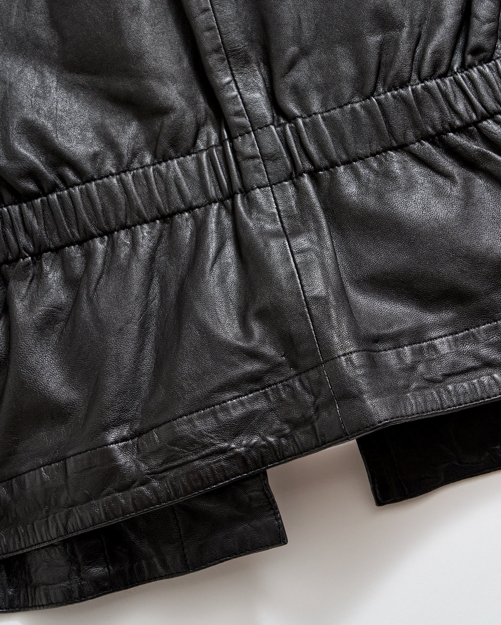 Close-up of a black leather garment, showing gathered and stitched detailing along the waist and hem areas, highlighting the texture and sheen of the material.