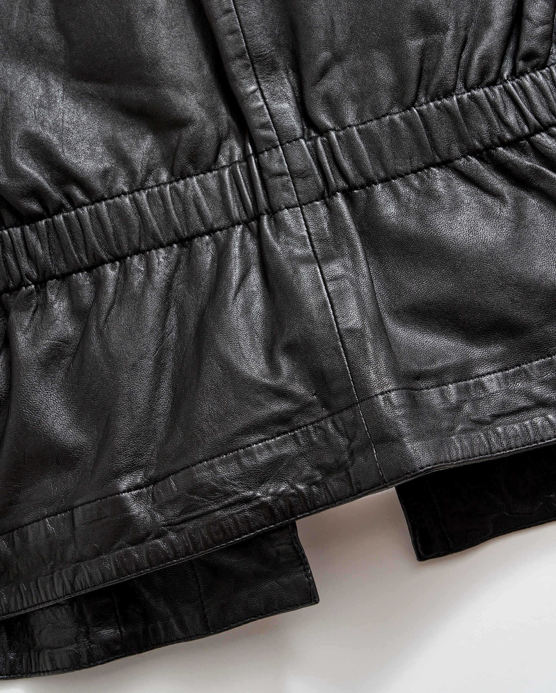 Close-up of a black leather garment, showing gathered and stitched detailing along the waist and hem areas, highlighting the texture and sheen of the material.