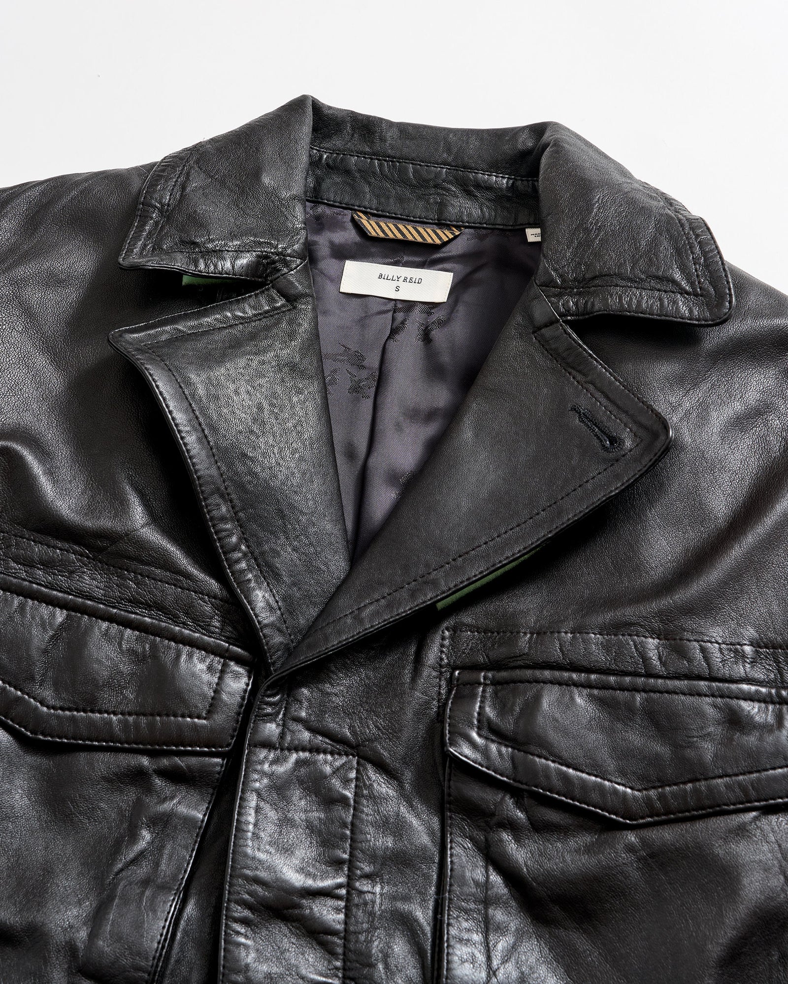 A close-up of a black leather jacket with a visible collar, front pockets, and a partially open front showing a tag labeled “BALLY KEAL” on the inside lining.