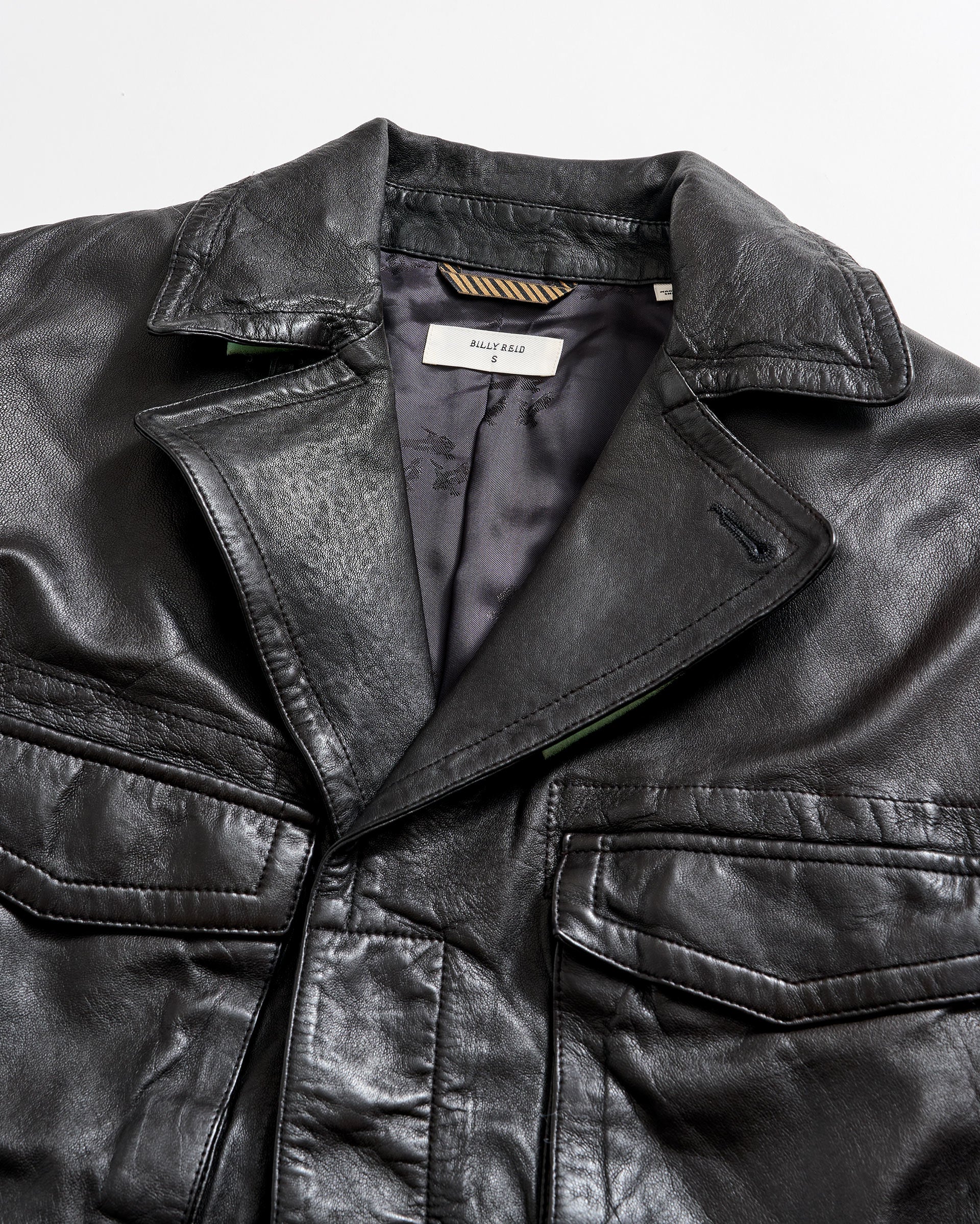 A close-up of a black leather jacket with a visible collar, front pockets, and a partially open front showing a tag labeled “BALLY KEAL” on the inside lining.