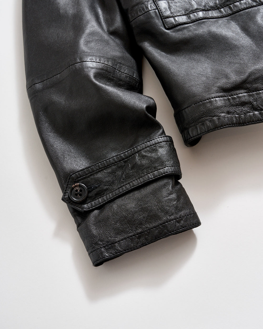 Close-up of the sleeve of a black leather jacket with a visible buttoned strap detail near the cuff, shown against a light background.