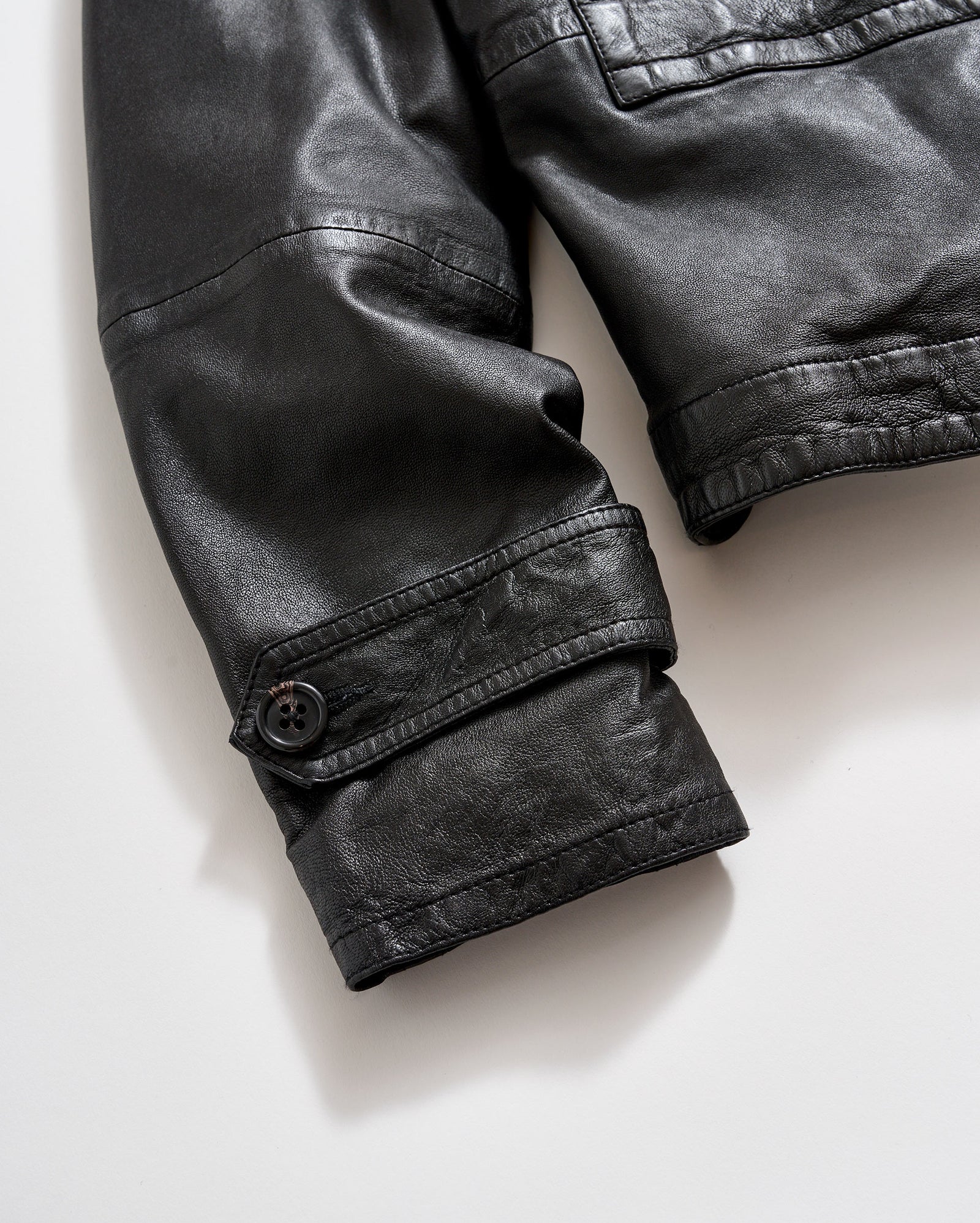 Close-up of the sleeve of a black leather jacket with a visible buttoned strap detail near the cuff, shown against a light background.