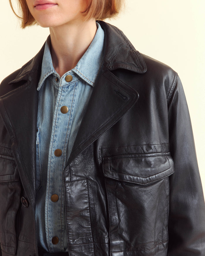 A woman with a light brown bob haircut wears a light blue denim shirt with metal buttons under a black leather jacket. The image is cropped to show her from the shoulders to the lips.