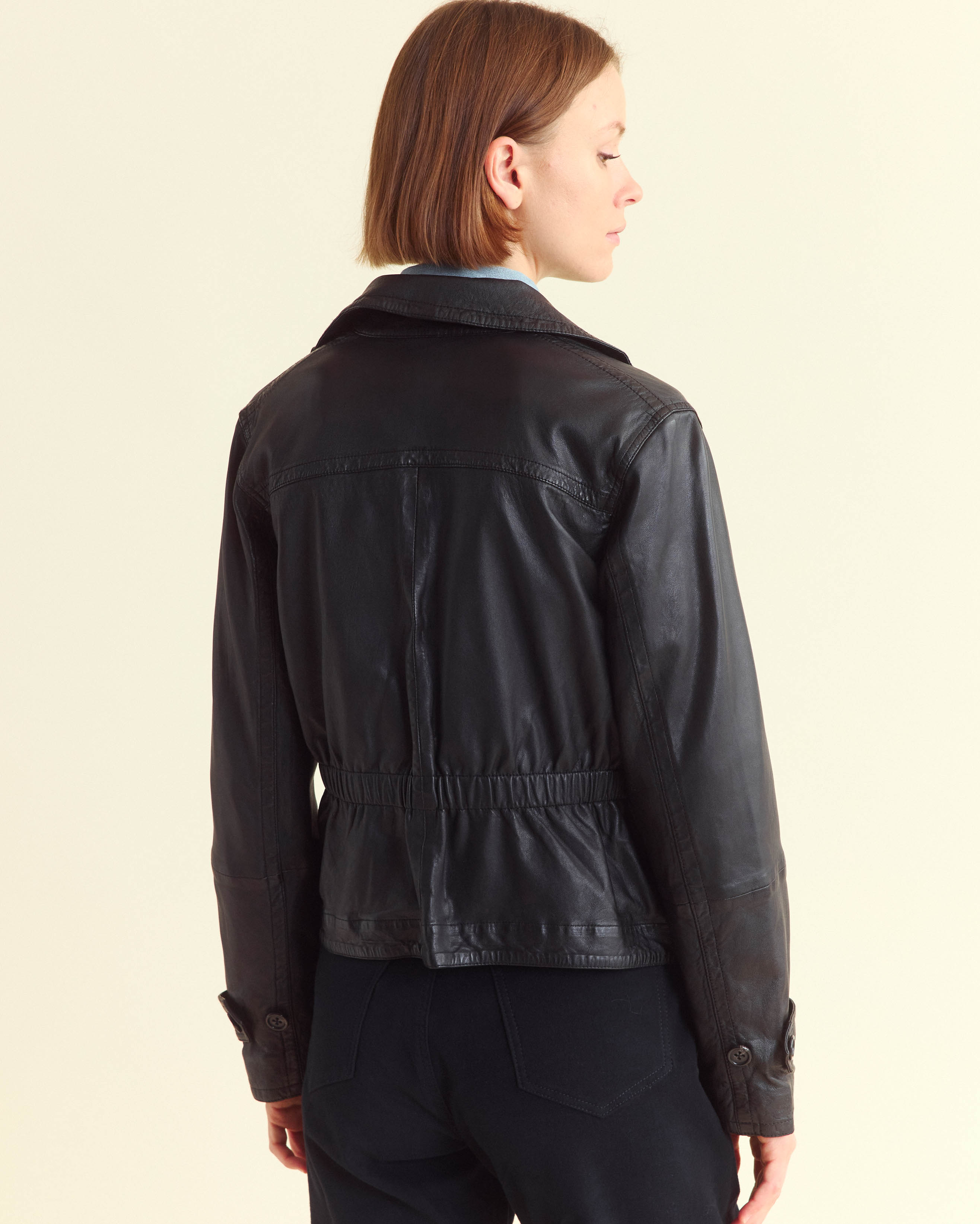 A person with short brown hair is standing and facing away from the camera, wearing a black leather jacket and dark pants against a plain light-colored background.