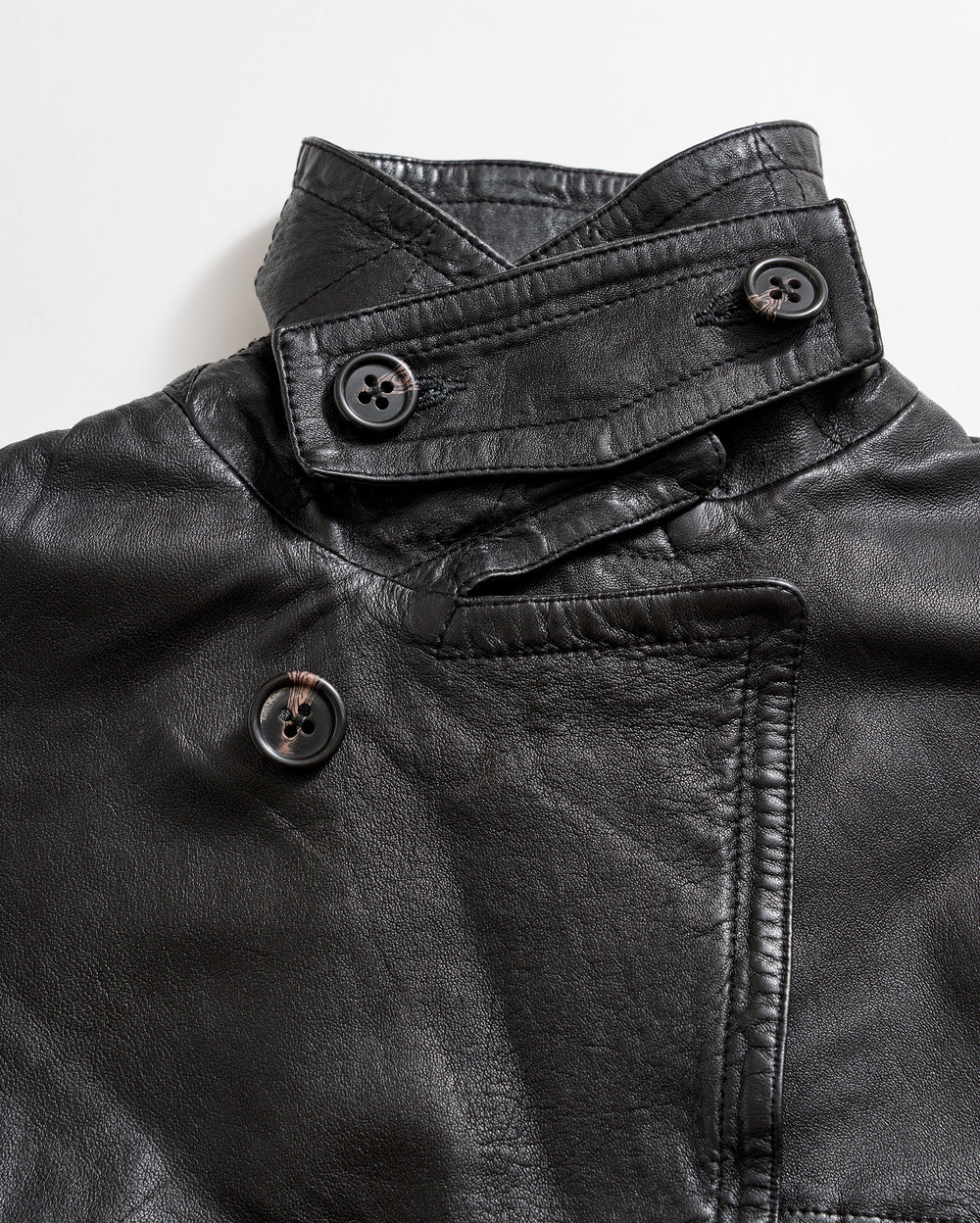 Close-up of a black leather jacket featuring an asymmetrical collar and three large black buttons, two on the collar and one on the front, against a white background.
