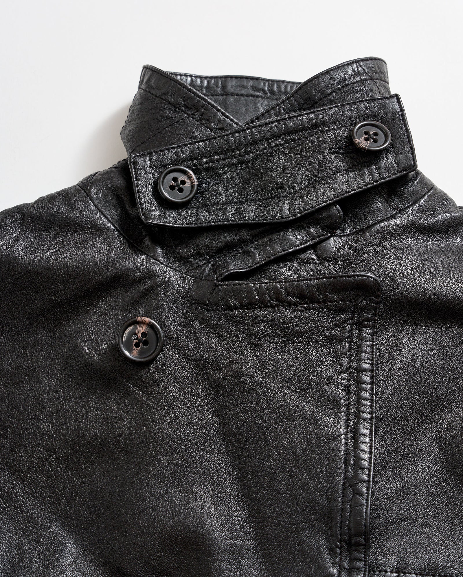 Close-up of a black leather jacket featuring an asymmetrical collar and three large black buttons, two on the collar and one on the front, against a white background.