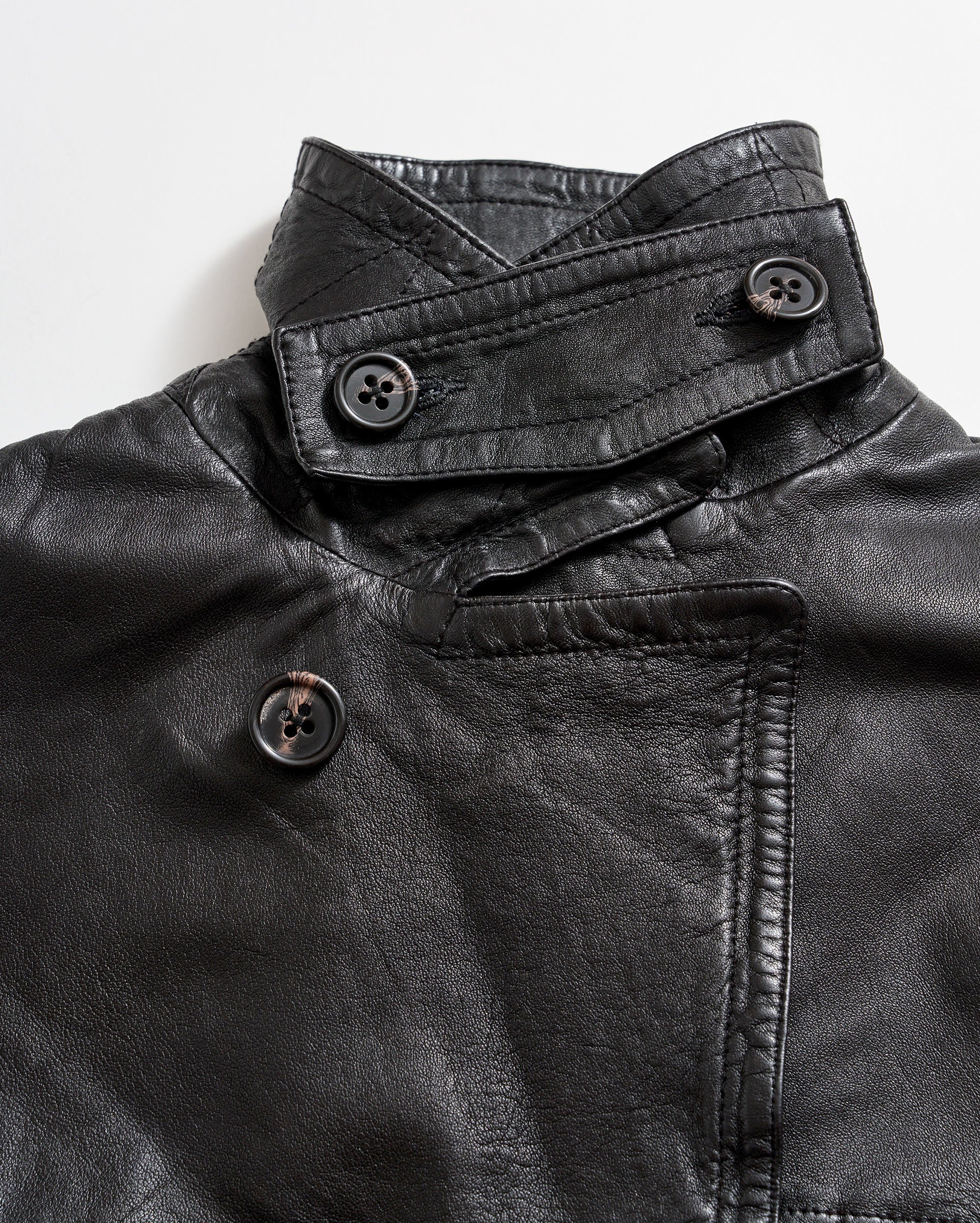 Close-up of a black leather jacket featuring an asymmetrical collar and three large black buttons, two on the collar and one on the front, against a white background.