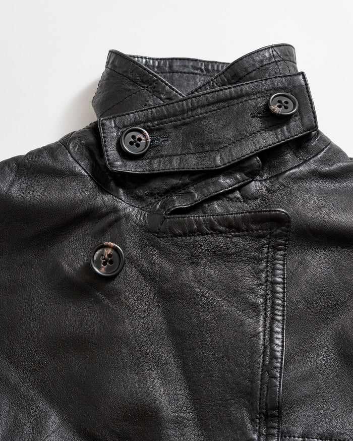 Close-up of a black leather jacket featuring an asymmetrical collar and three large black buttons, two on the collar and one on the front, against a white background.
