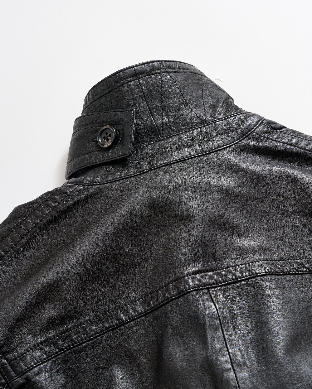 Close-up of the upper back and collar of a black leather jacket with visible stitching and a buttoned strap detail on the collar, set against a plain white background.