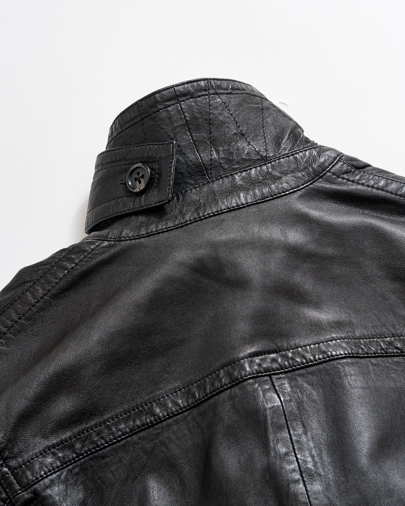 Close-up of the upper back and collar of a black leather jacket with visible stitching and a buttoned strap detail on the collar, set against a plain white background.