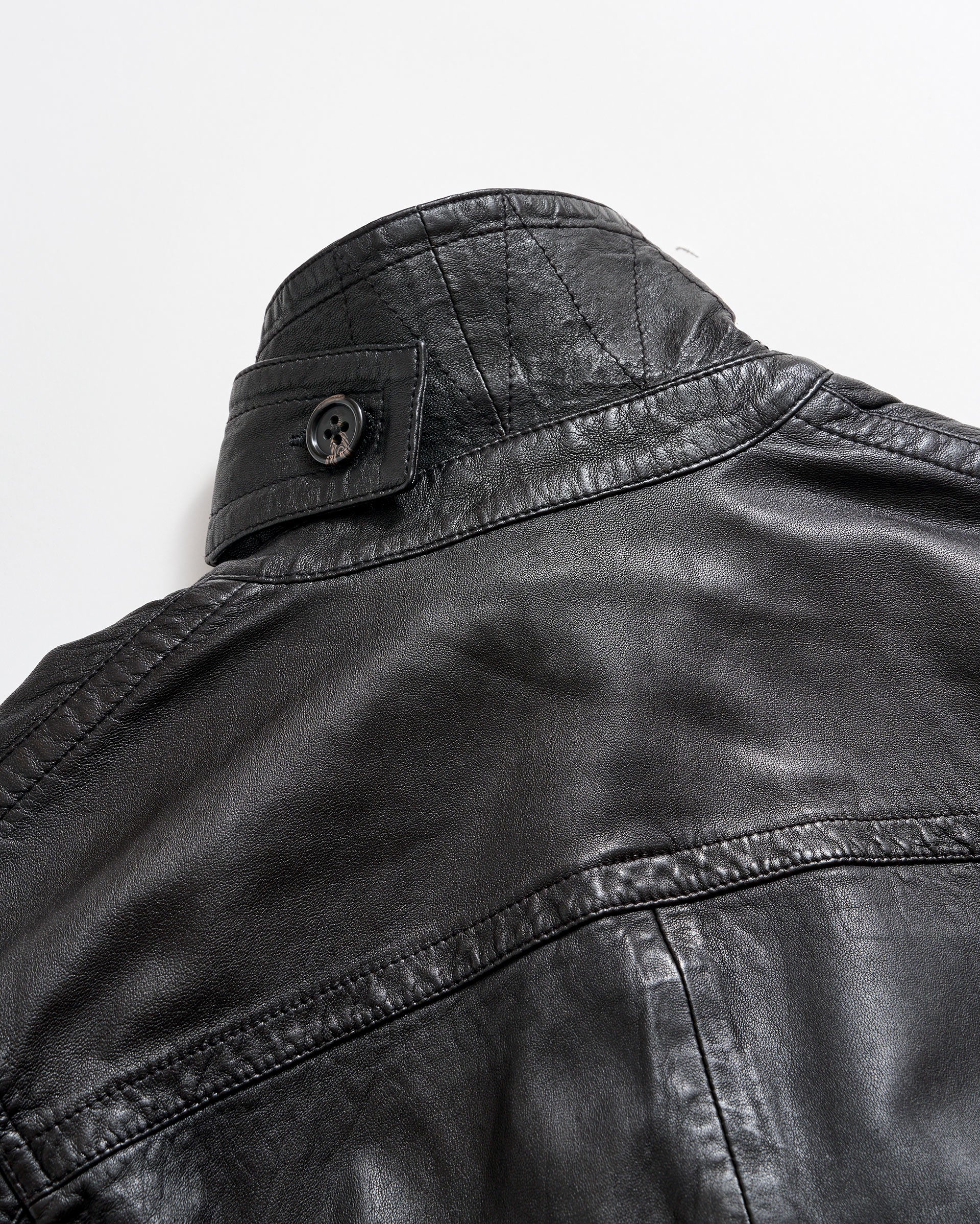 Close-up of the upper back and collar of a black leather jacket with visible stitching and a buttoned strap detail on the collar, set against a plain white background.