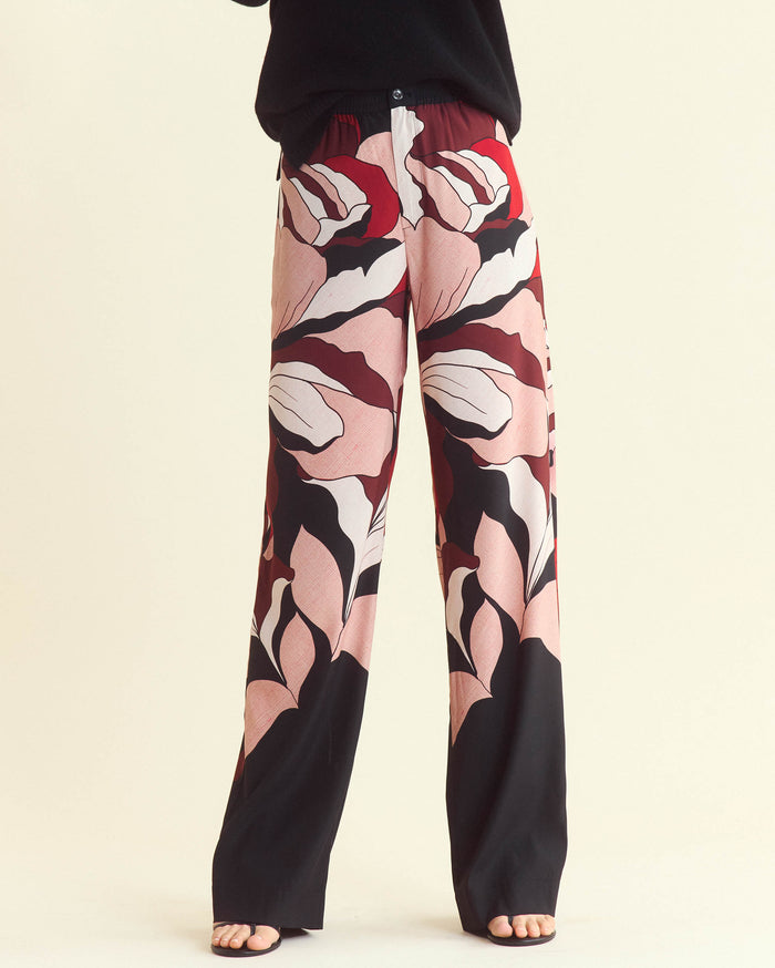 Person wearing wide-leg pants with a bold, abstract pink, red, black, and white floral pattern. Only the lower half of the body is visible; feet are in sandals and the background is light beige.