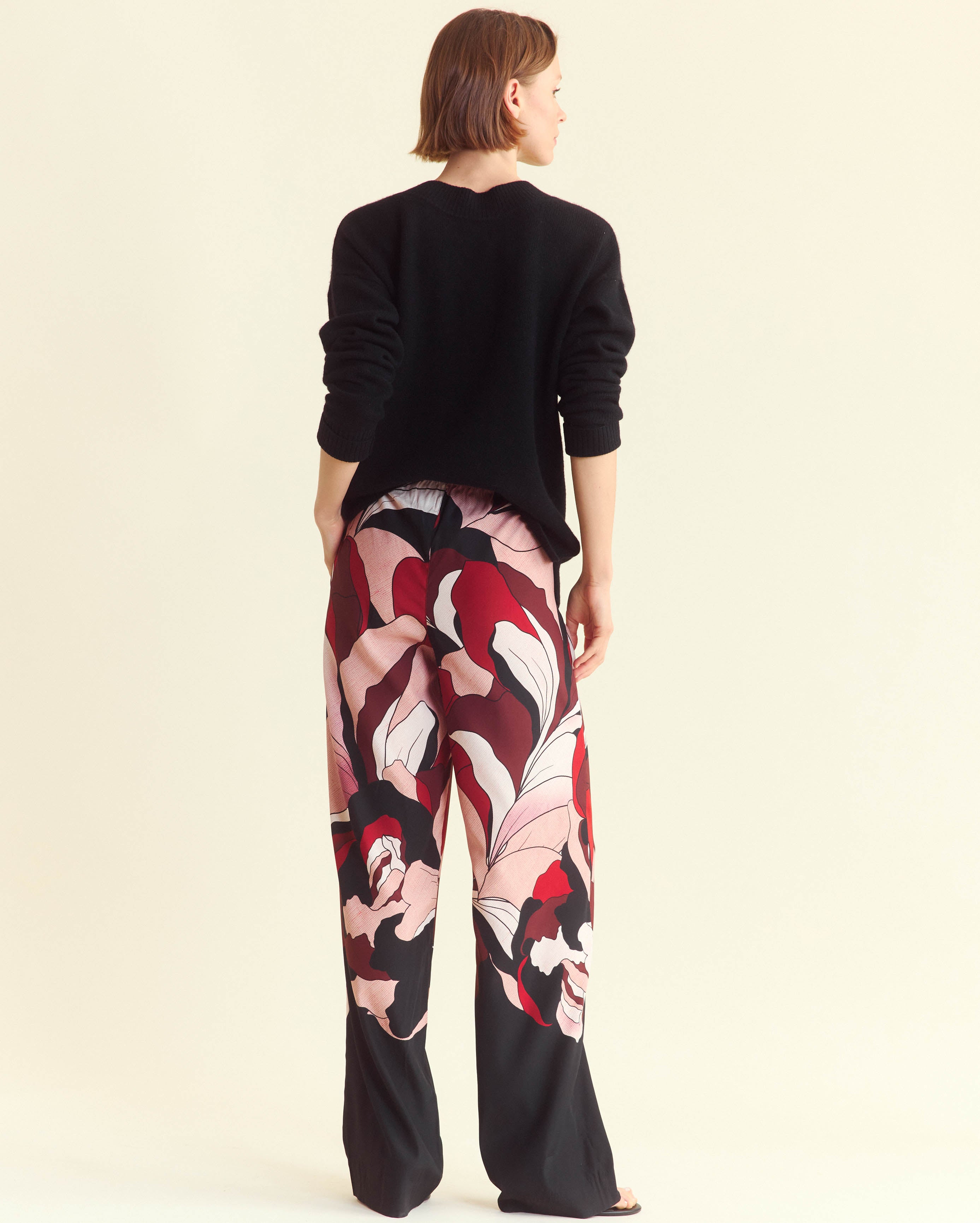 A person stands facing away, wearing a black long-sleeve top and loose, wide-leg pants with a bold, abstract floral pattern in red, pink, white, and black. The background is a plain off-white color.