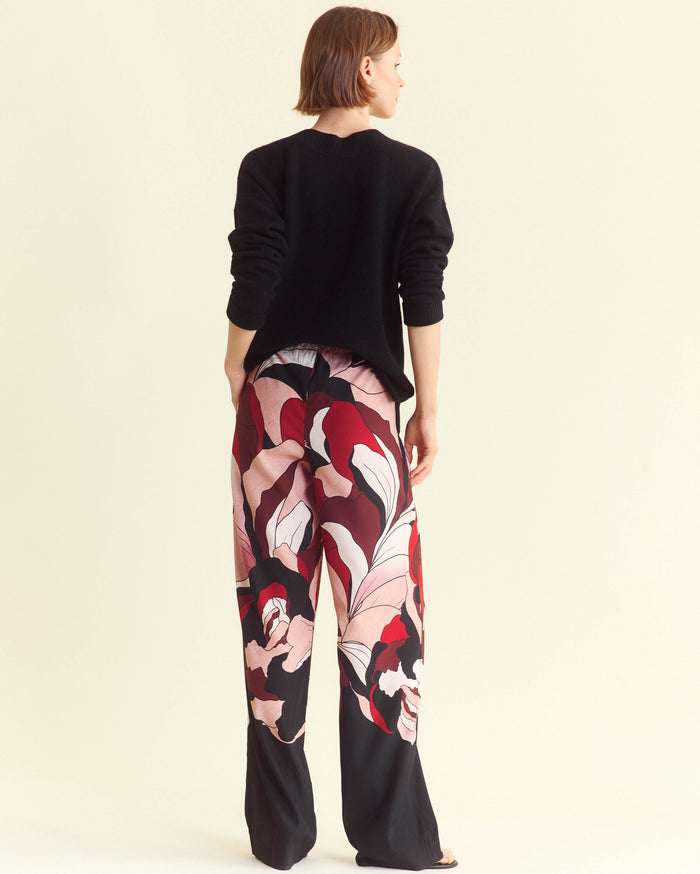 A person stands facing away, wearing a black long-sleeve top and loose, wide-leg pants with a bold, abstract floral pattern in red, pink, white, and black. The background is a plain off-white color.