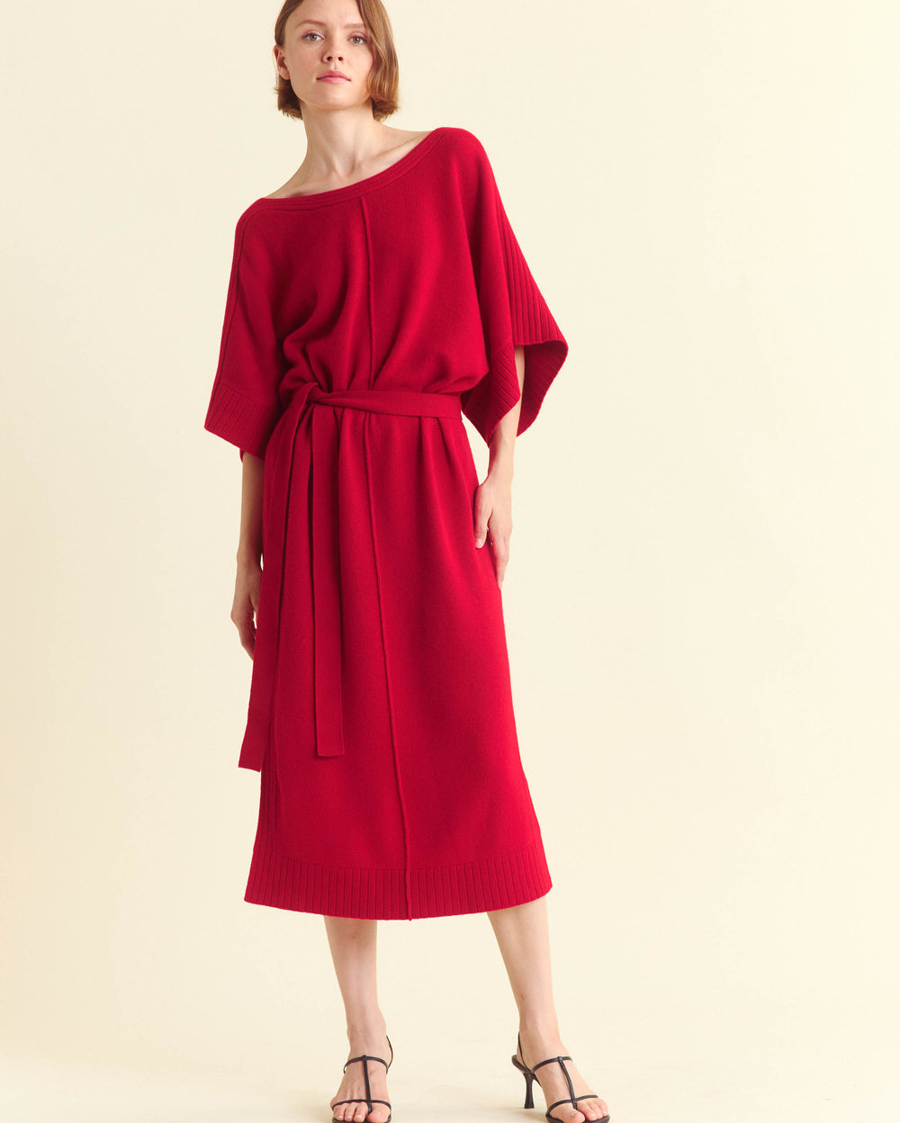 A woman wearing a loose-fitting, bright red midi dress with wide sleeves and a matching belt stands against a plain, light background. She is wearing black strappy sandals and has a neutral expression.